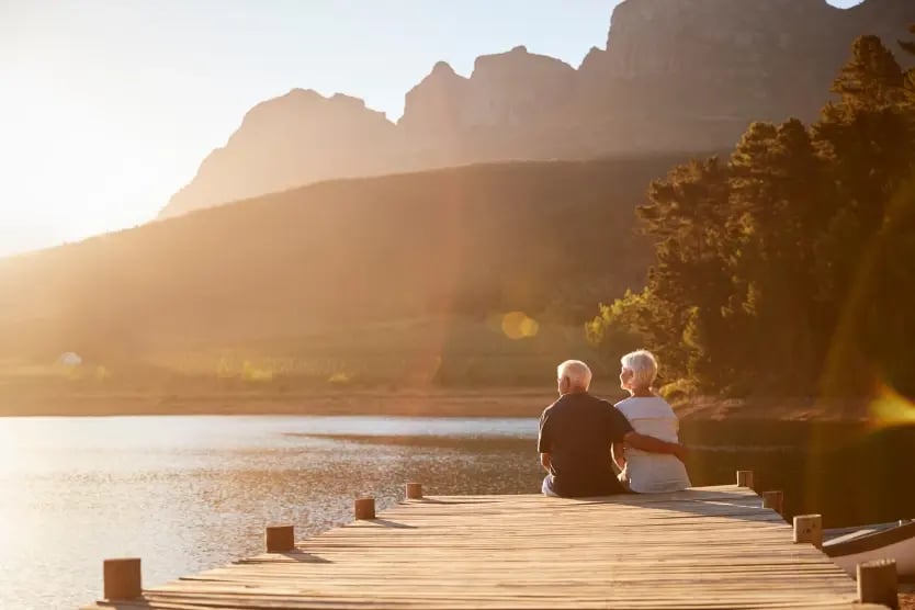 Older, retired couple sitting on a dock while enjoying a beautiful lake scene.
